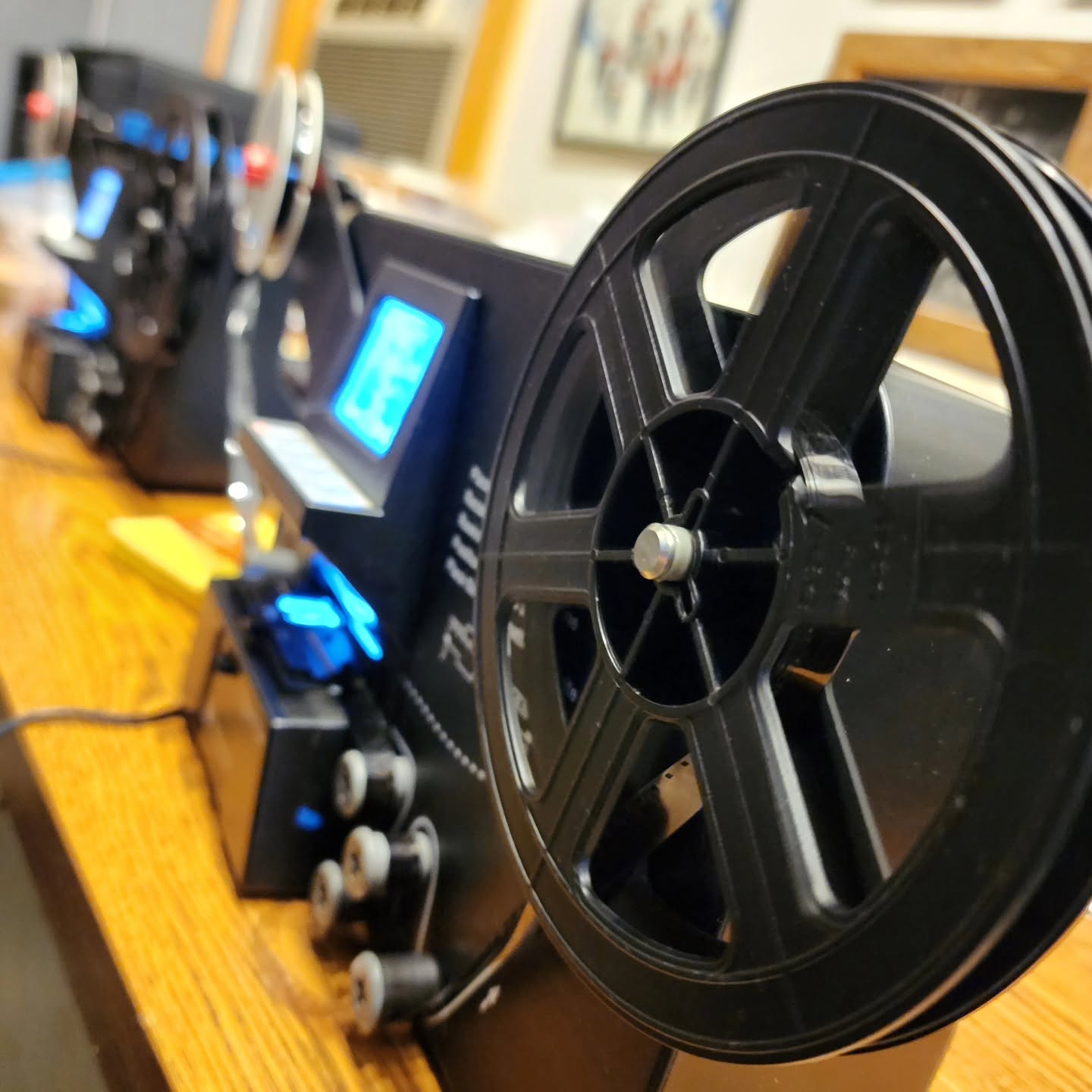 Close-up of a black film projector reel mounted on a desk, with a blue LCD display visible in the background.